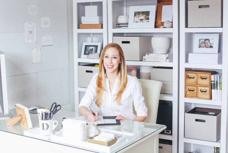 A woman sits smiling at a white desk. The office is light with styled white shelves in the background