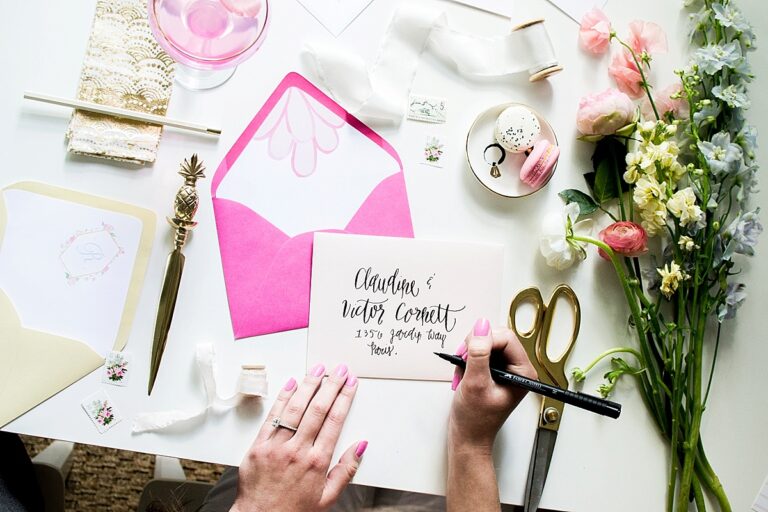 A flat lay of stationary and decorative items with a woman's hand writing calligraphy on a card.