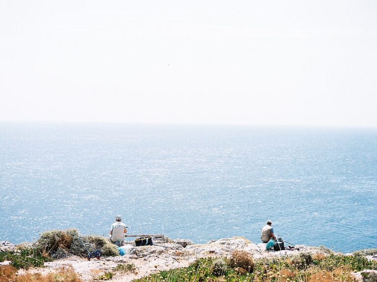 Two men sit on a cliffside, fishing into a large expanse of water.