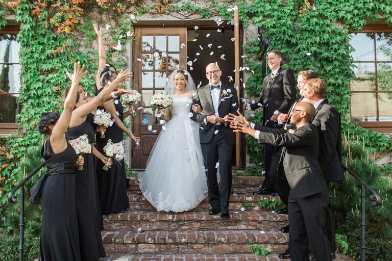 A wedding party throws petals in the air as a newlywed couple exits a wedding venue.