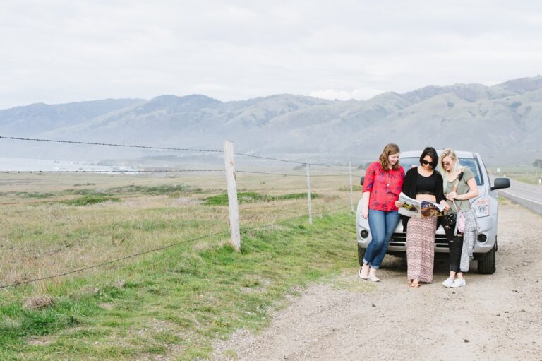 Three women stand in front of a car on the side of a road, reading from a magazine. In the distance are mountains.