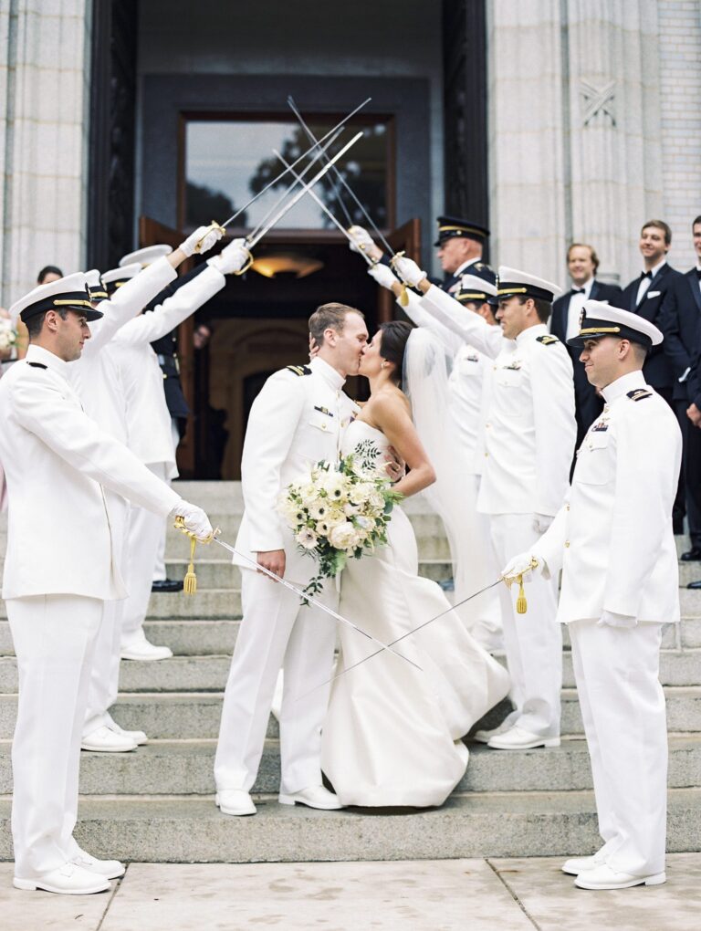 A newly married couple in all white kiss on the external stairs of a building underneath ceremonial swords held in the air by military members dressed in white