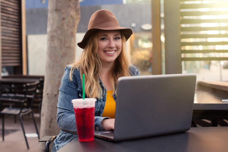 A woman works outside at a laptop with an iced drink, smiling at the camera.