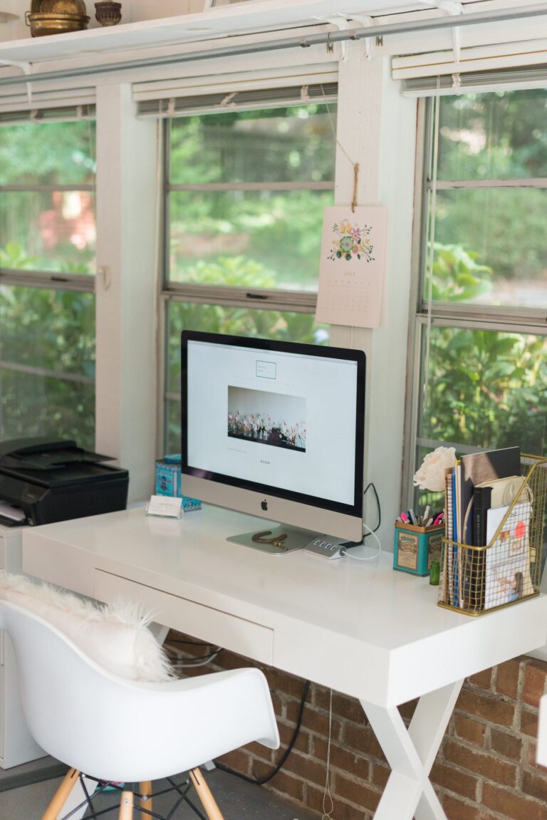 A white desk with an iMac in font of a wall of windows showing greenery outside.
