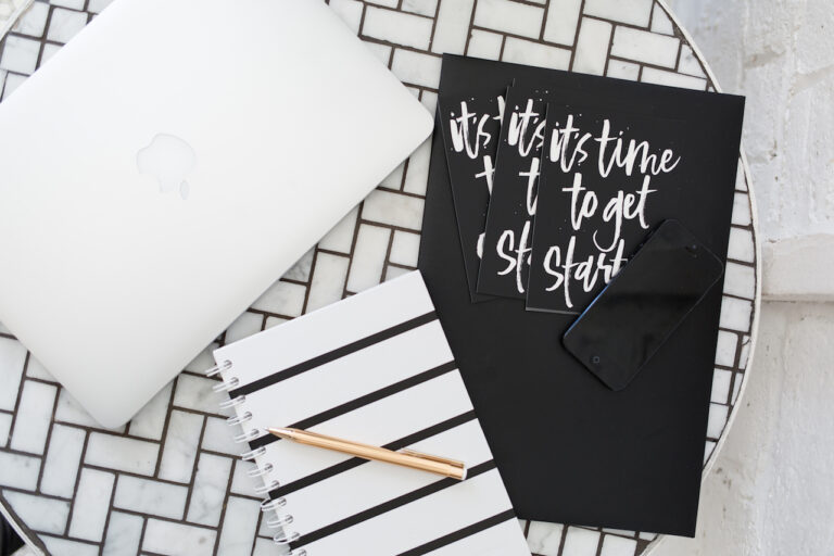 A white mosaic tile table with black grout is set with an iMac, a notebook, and folder with cards.