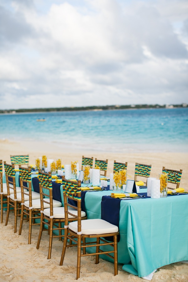 A long dining table and chairs on the beach, decorated for guests.