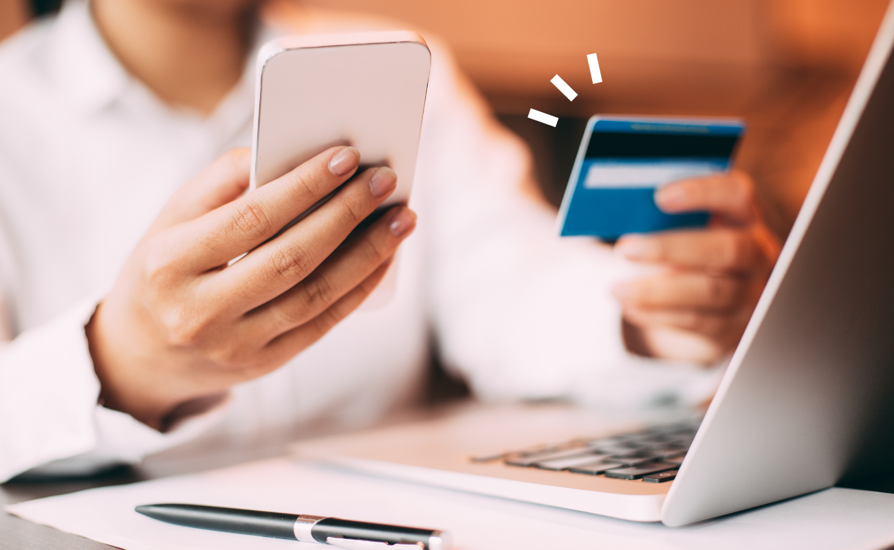 Close up of someone's hands holding a phone in one and a credit card in another with a laptop open in front of them