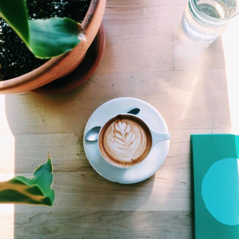 A coffee with latte art sits on a table framed by plants and assorted objects