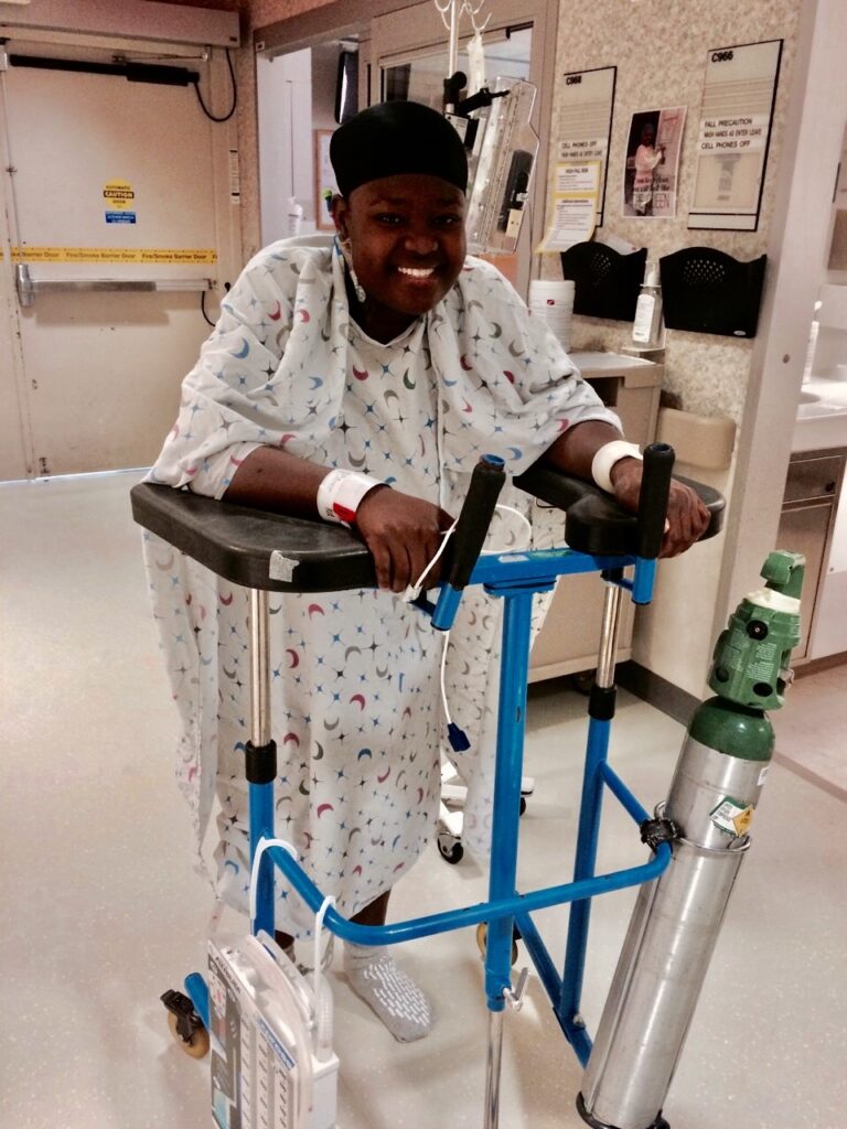 A woman in a hospital gown smiles as she leans on an assisted walking device with medical equipment