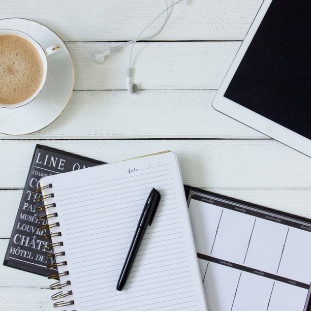 A notebook, desk calendar, cup of coffee, tablet, and earbuds on a slatted wooden surface.