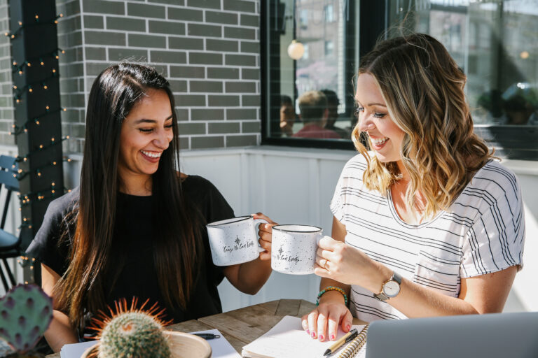 Two women cheers with coffee mugs as they sit outside.