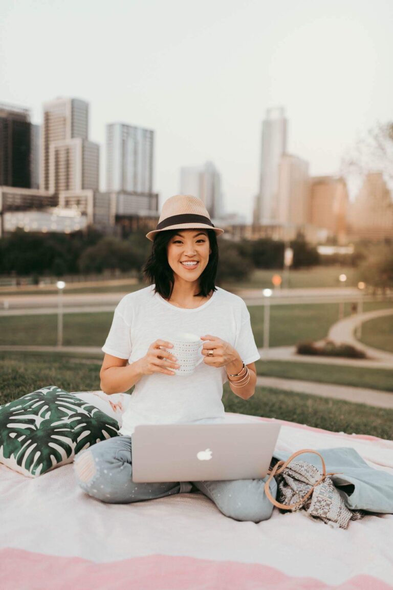 A woman sits cross-legged on a picnic blanket in front of a city skyline. She holds a mug in her hands on a laptop on her legs.