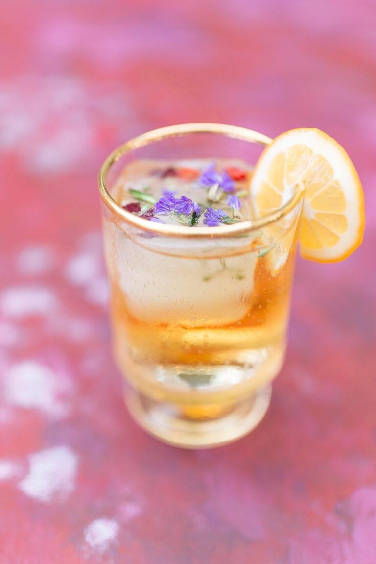 A drink with a single, large ice cube is garnished with a slice of lemon and flower petals sits on a pink surface.