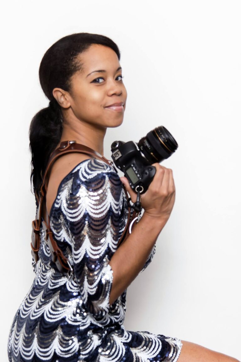 A woman in a sequined dress poses with a camera in front of a white wall