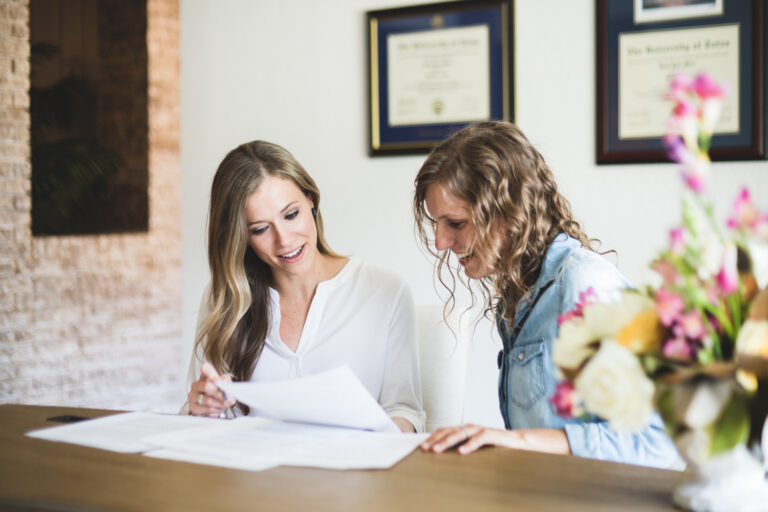 Two women sitting at a counter, glancing down at a group of papers and smiling.