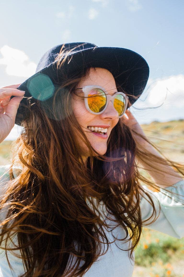 Close up of a woman outside in the sun, smiling. She wears sunglasses and a brimmed hat