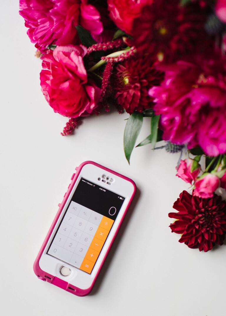 A phone with a pink case sits on a white surface with a calculator app open on the screen. The phone is framed by pink flowers.