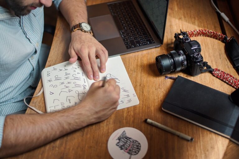 a man writing in a notebook with a laptop and camera on his desk