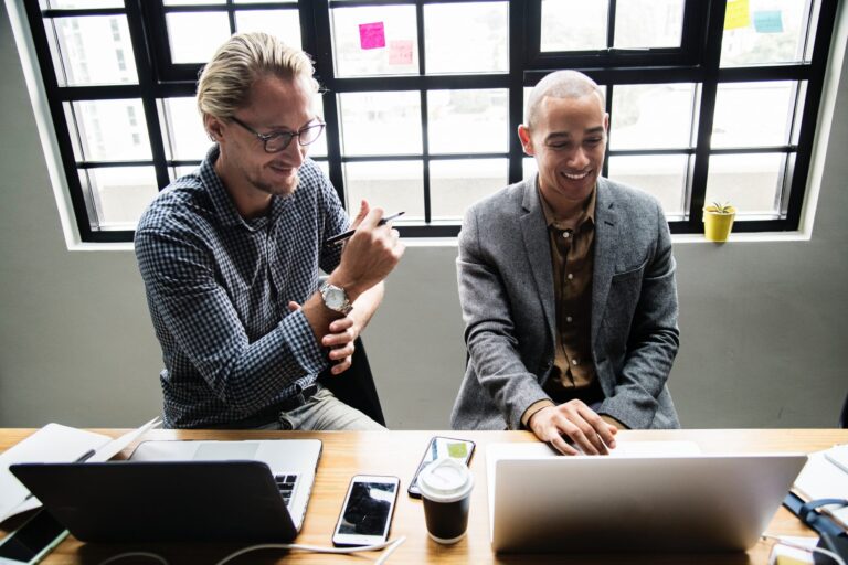 Two men work at a desk in front of a large window, smiling at a laptop