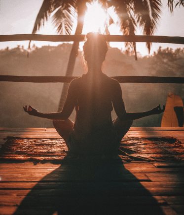 A woman, backlit by the sun, meditates in front of a palm tree