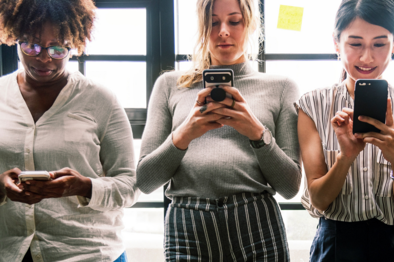 Three women stand side by side in front of windows, looking at their phones