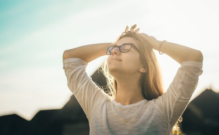 A woman wearing glasses basks in the sun with hands on her head