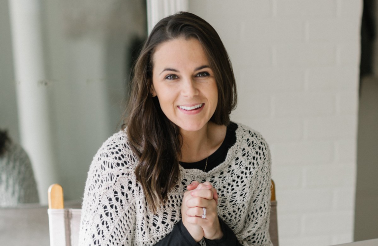 A headshot of a woman sitting in front of a white wall and smiling at the camera.