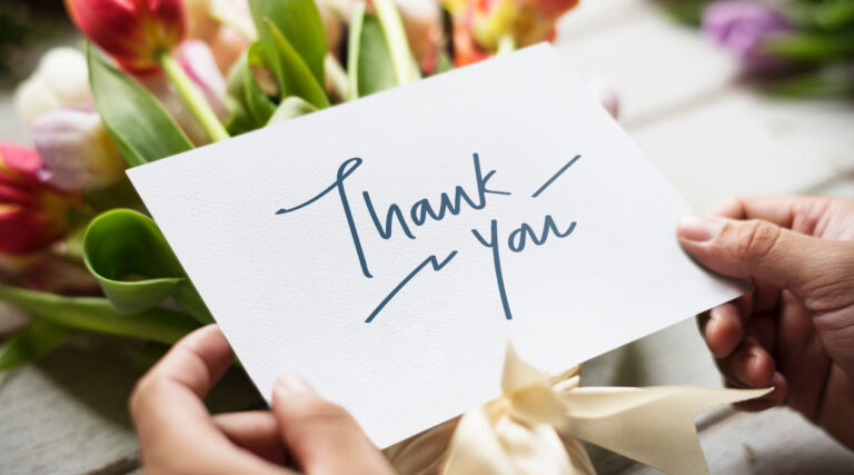A pair of hands hold a thank you card in front of a bouquet of flowers