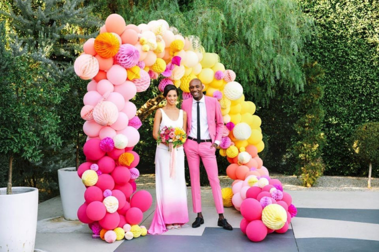 Bride and Groom in bright pink formal wear smile together under balloon arch.