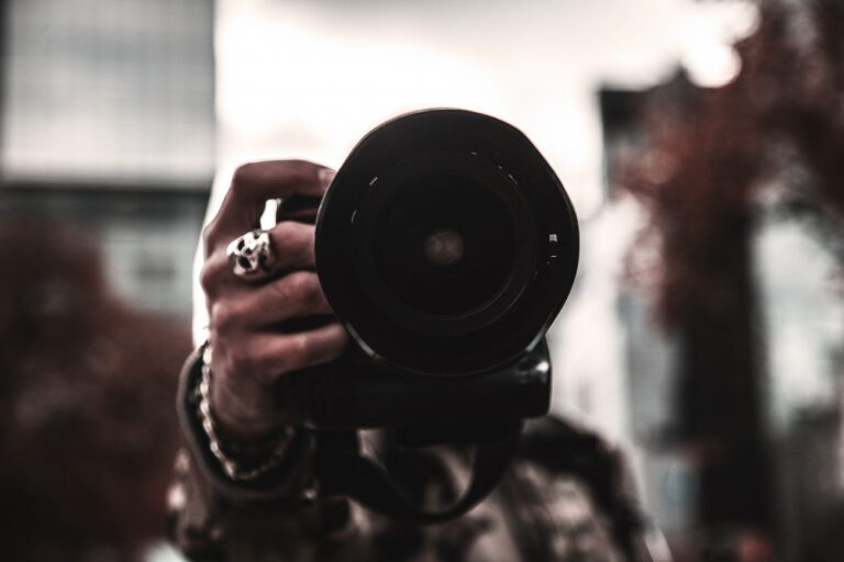 a hand with rings and a bracelet holding a camera with a large lens