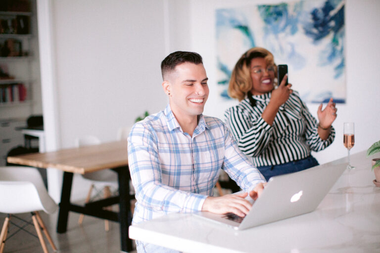 A man smiles, typing at a laptop. Beside him, a woman takes a picture on her phone