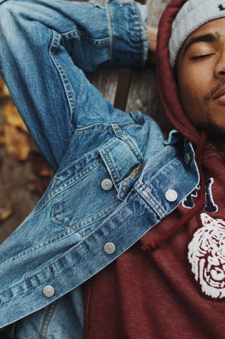 Close up of a man sleeping outdoors. He wears a maroon hoodie and denim jacket.