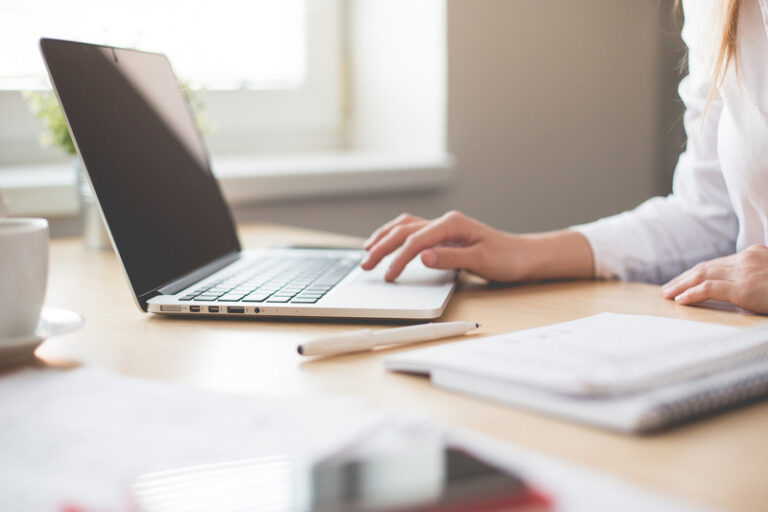 A person works at an open laptop on a desk with notebooks and coffee cup