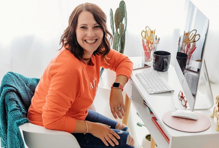 Woman in an orange sweater at desk smiles over her shoulder.