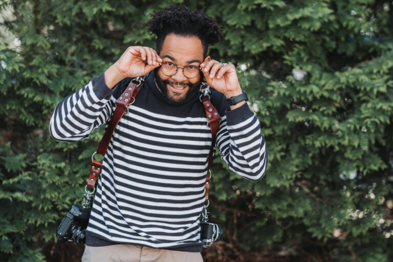 A man stands in front of shrubbery, wearing camera equipment, and smiling at the camera.