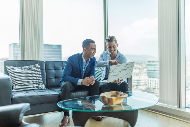 Two men in suits smile at a laptop in front of windows with a city view.