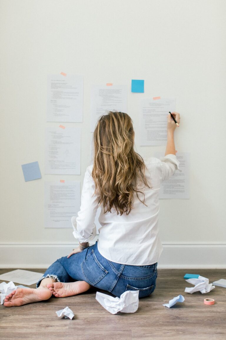 A woman sits on the floor in front of a wall, facing away from camera. She writes on pieces of paper taped to the wall.