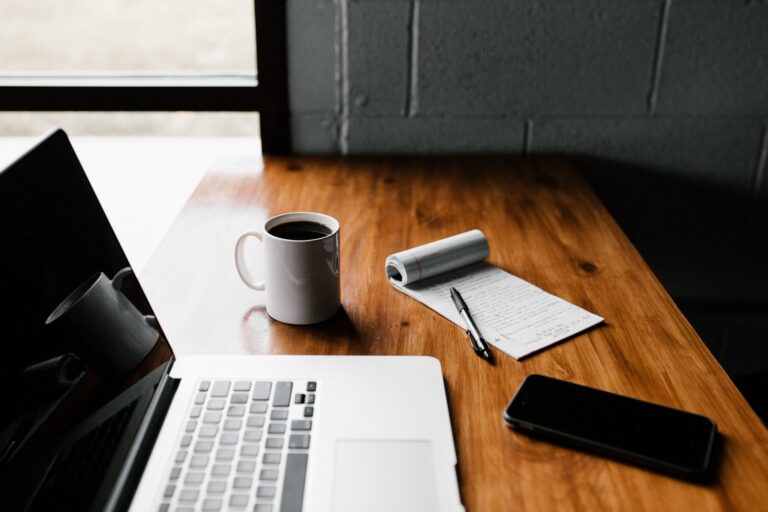 A laptop sits open on a wooden desk with a mug of coffee, notepad, and phone