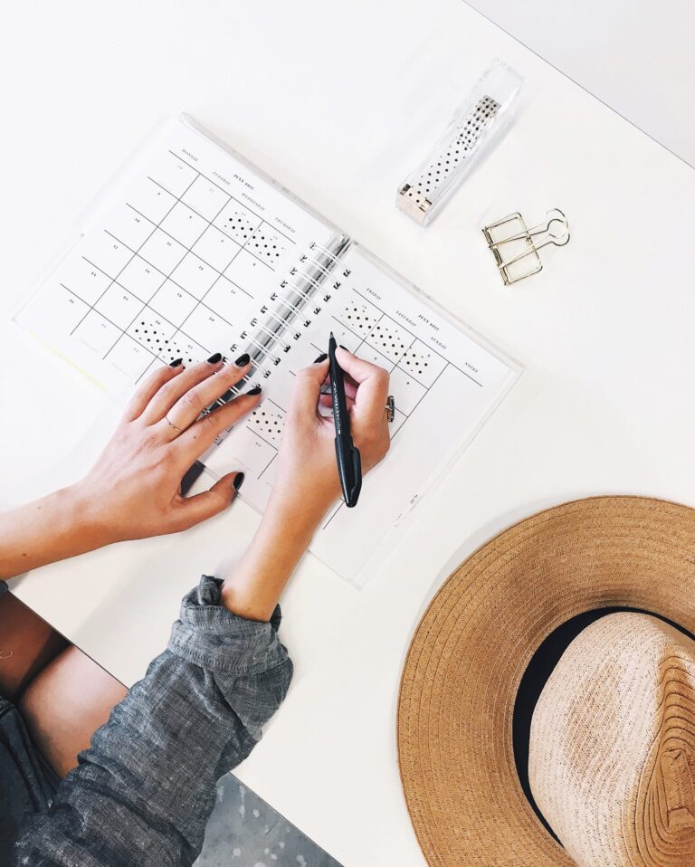 Close up of a woman's hands writing in a planner while a straw hat rests on the table.