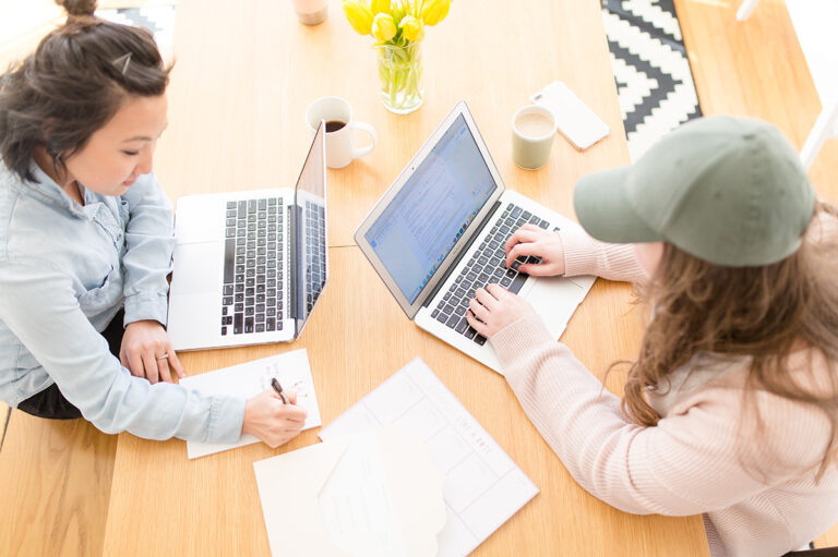 Aerial view of two women working on laptops at a wooden table.