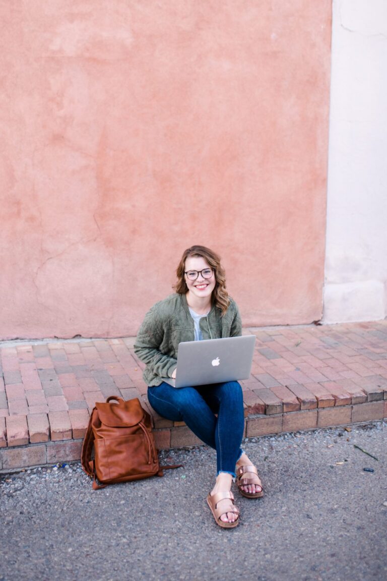 A woman sits on a brick curb, working on her laptop and smiling at the camera