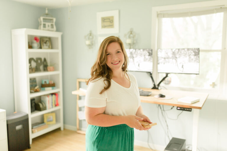 A woman stands in the middle of a home office, holding a calculator and smiling at the camera