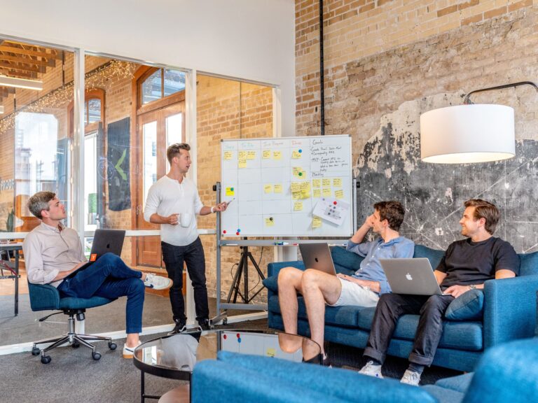 A man stands next to a whiteboard, giving a presentation to three men working on laptops.