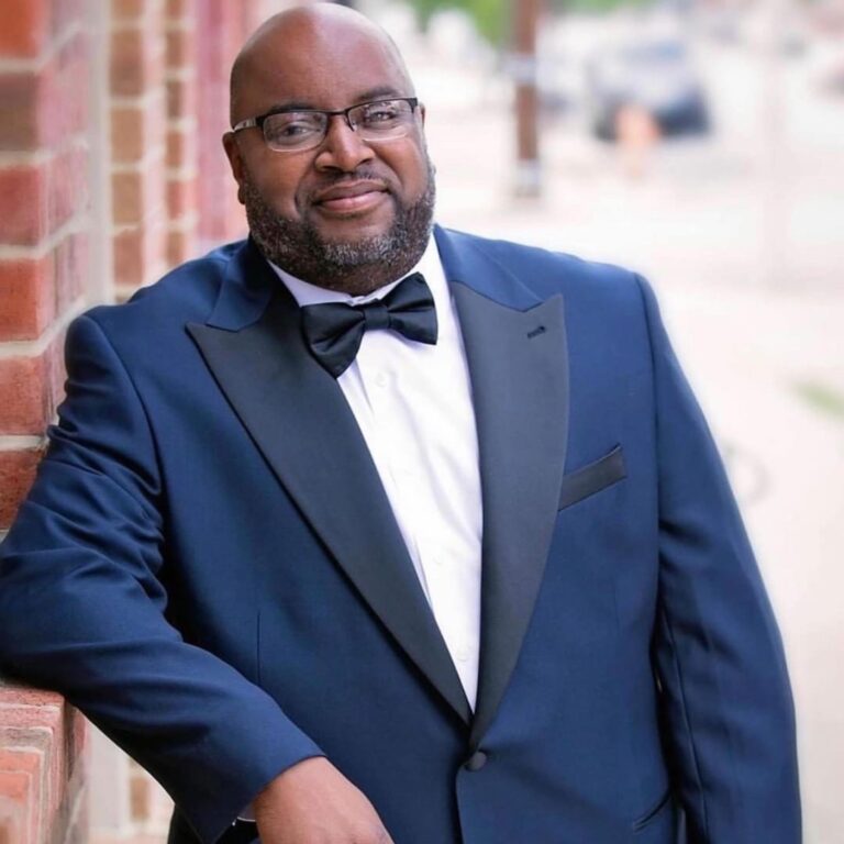 A headshot of a man in a blue suit and black bowtie, leaning against a brick wall