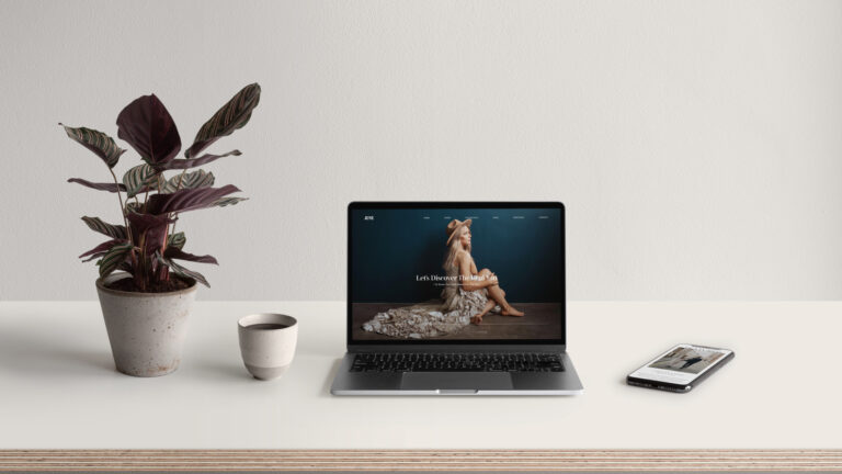 A laptop sits open with an image of a woman as the wallpaper. Beside the laptop are a cell phone, a ceramic cup, and a potted plant.