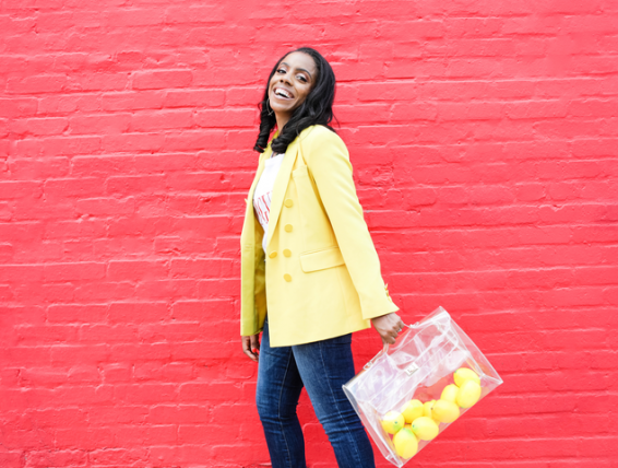 a smiling woman in a yellow blazer in front of a red brick wall carrying lemons