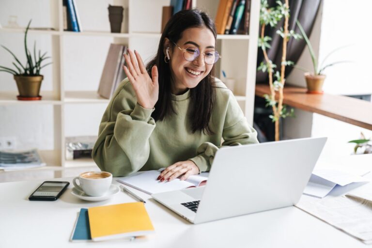 a woman with glasses sitting at a desk waving to her laptop