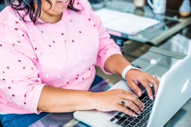 A woman in a pink sweater works at a laptop on a glass desk.