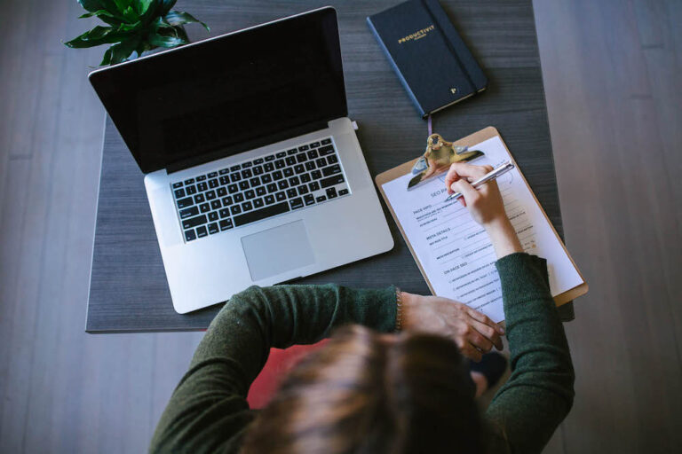 Aerial view of a person working at a table, writing on a clipboard. Beside them is an open laptop