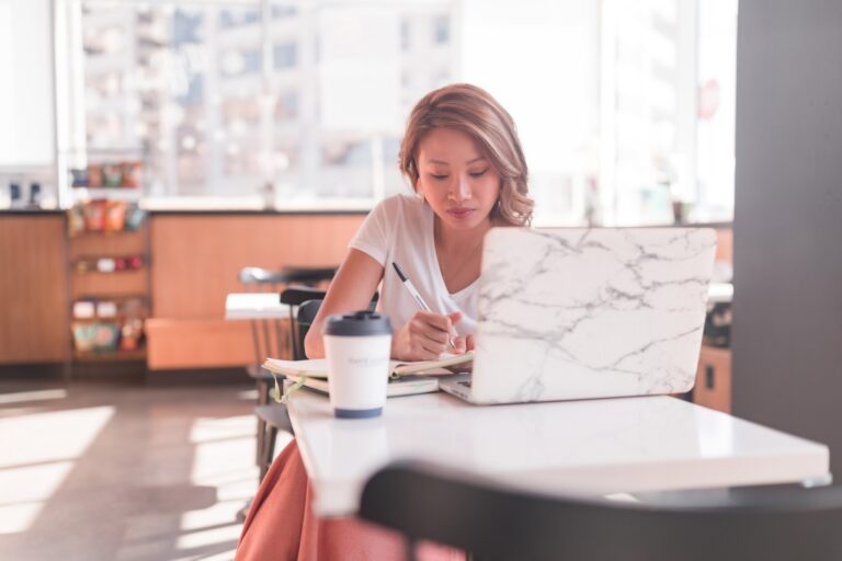 A woman sits at a table in a coffee shop, working on her laptop and writing in a notebook